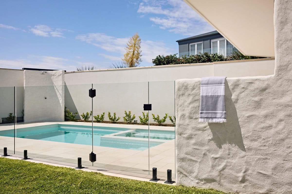 The pool and spa seen across the lawn through frameless glass fencing, a striped towel hanging on the rendered wall.