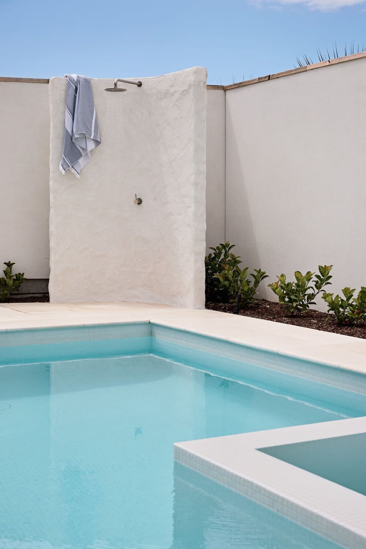 An outdoor shower set into a sculpted white rendered blade, with a towel over the wall and the pool below.