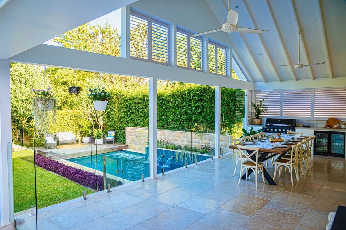 A covered outdoor pavilion with vaulted ceiling, long dining table and built-in barbecue, looking out over the pool.