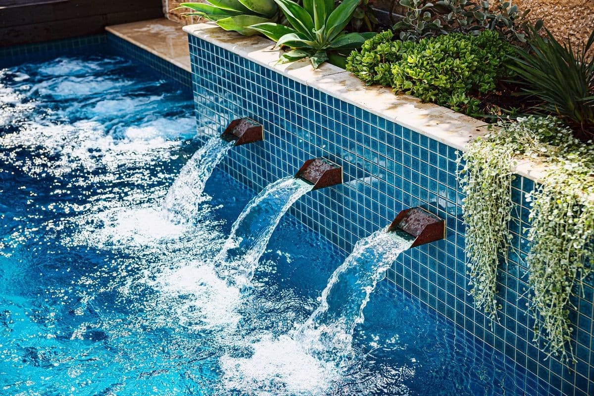 Three copper water blades arcing from the blue-tiled planter wall into the pool, with trailing greenery above.