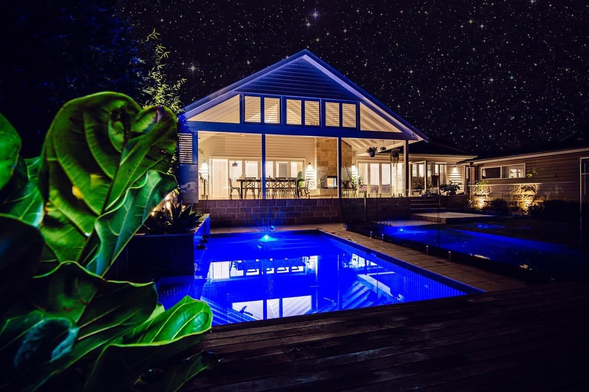 The pool and pavilion lit at night under a star-filled sky, the water glowing blue against the warm interior.