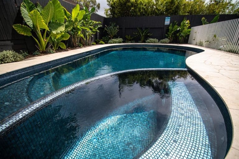 The pool and spa from the paved terrace, bananas and strelitzia along the black boundary fence.