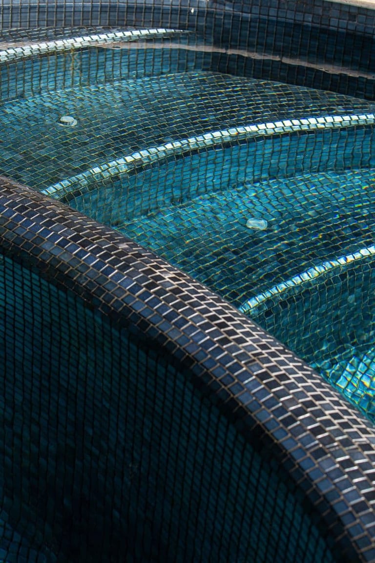 The spa steps in iridescent mosaic, water spilling over the curved edge into the pool.