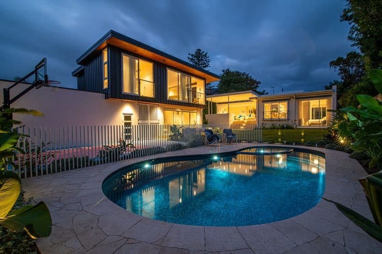 The pool lit at night, the two-storey house glowing warm behind the crazy-paved terrace.