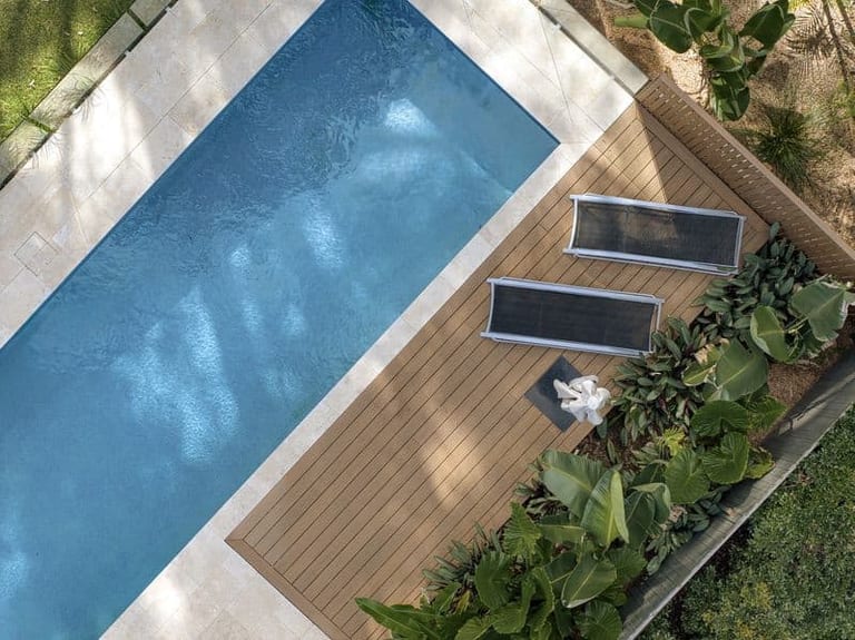 Aerial detail of the pool meeting the timber deck, loungers and a border of broad tropical foliage.