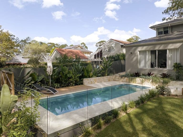The pool and glass fencing seen from the garden, with the rendered house and a sandstone planter behind.