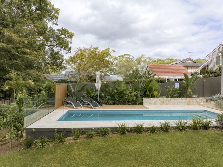 The pool and deck seen across the lawn, tropical planting and neighbouring rooflines beyond the fence.