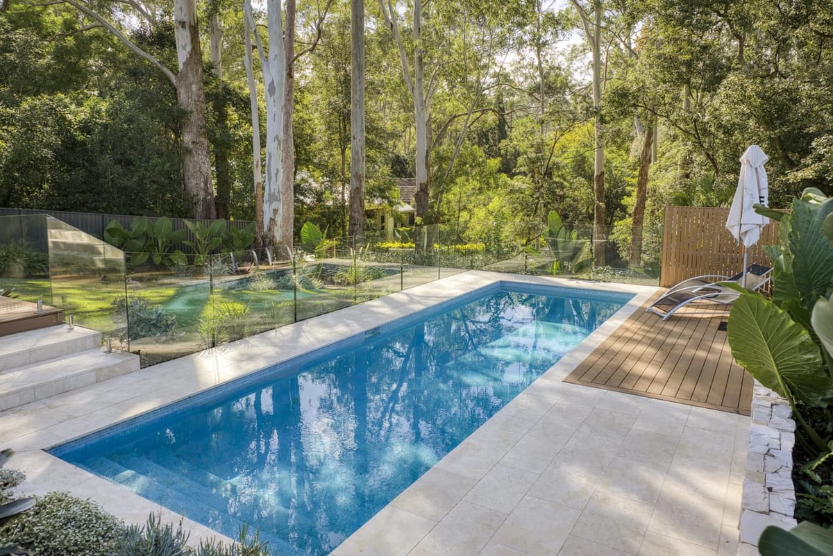 A long lap pool edged in travertine beneath tall gum trees, with a timber deck and lounger to one side and lawn beyond the glass fence.