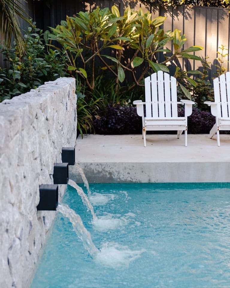Water arcing from a black spout in the pale stone wall, the chairs and planting behind.
