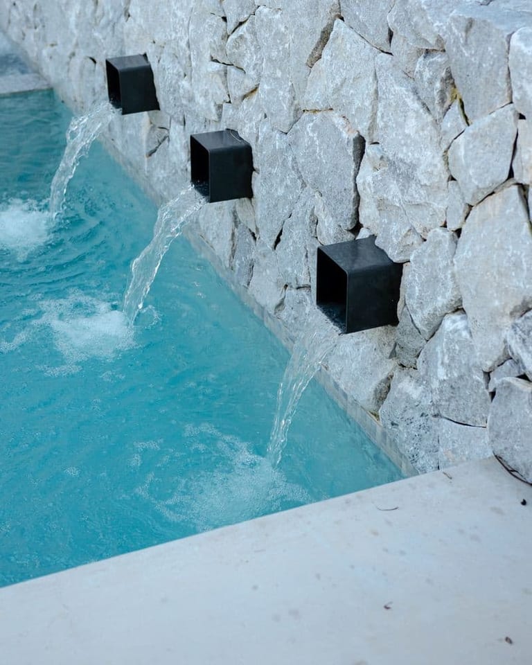 Three black spouts set into the rough stone wall, water falling into the pool below.