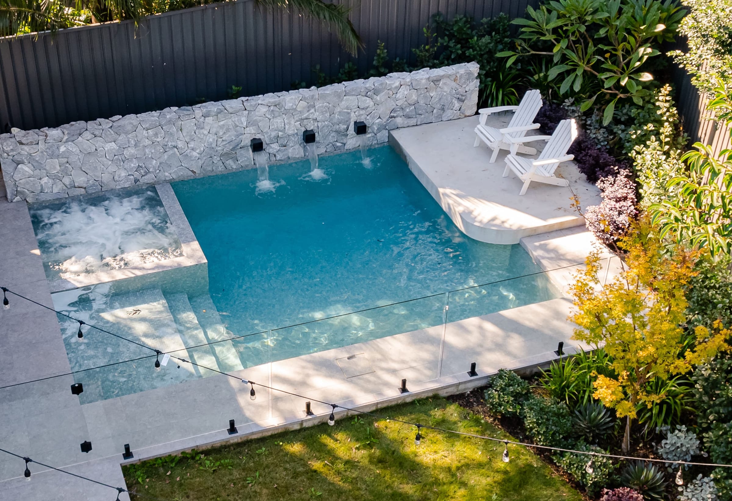 A pool with a cantilevered stone edge and a raised sun shelf, water spouting from three black outlets in a pale stone wall.