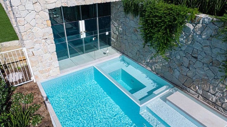 Elevated view of the shallow end, the spa and the glass-walled pavilion set into the stone wall behind it.