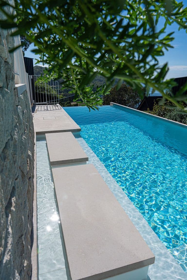 Stepping stones running beside the stone wall, with the pool and harbour view beyond the leaves.