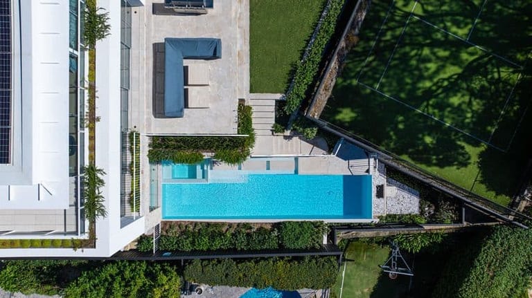 High aerial of the house, terrace and pool in its narrow terrace above the lawn and court.