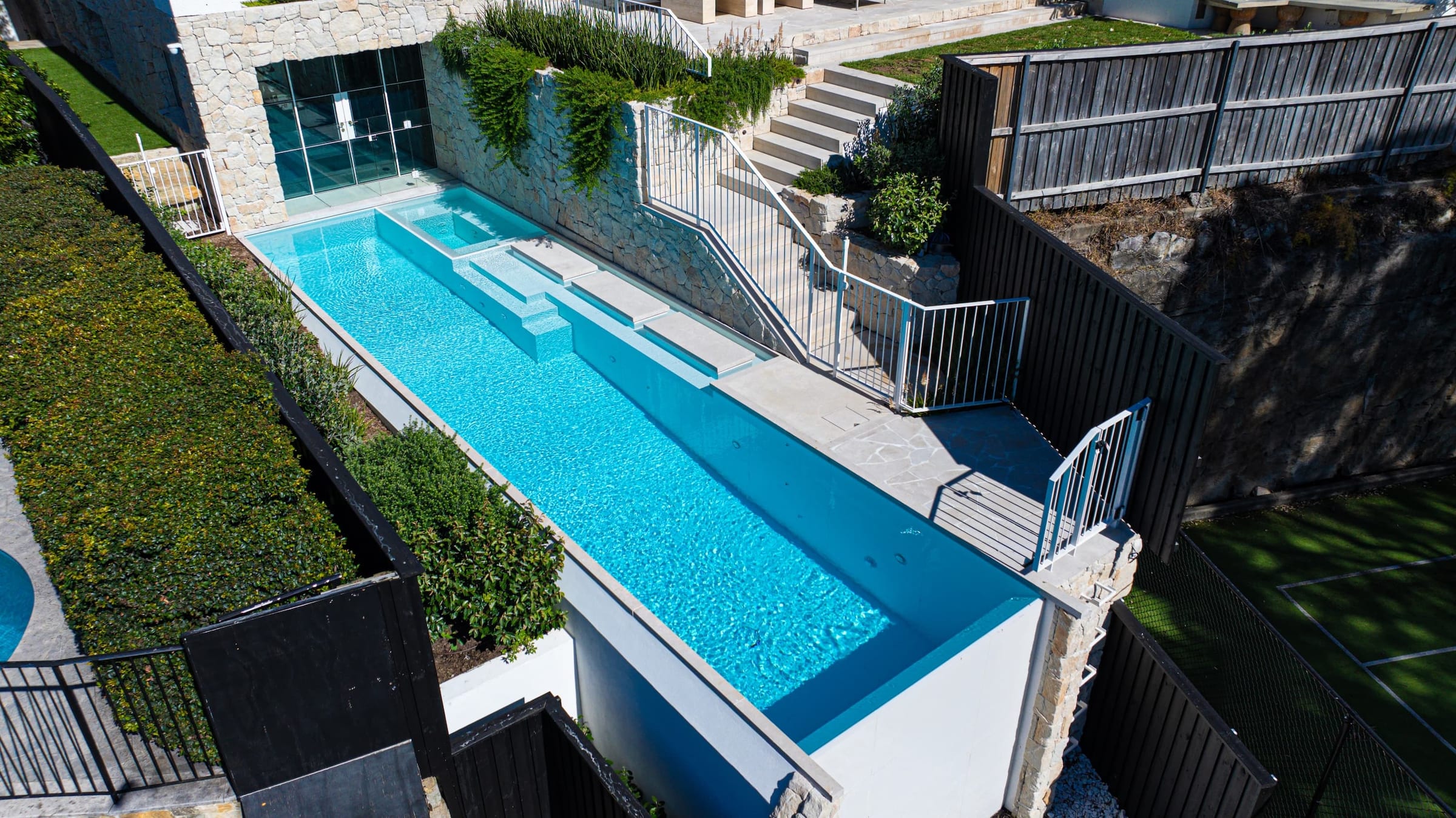 Aerial view of a twelve metre infinity-edge pool on a steep harbourside block, with stepping stones, a spa and a glazed pavilion at the far end.