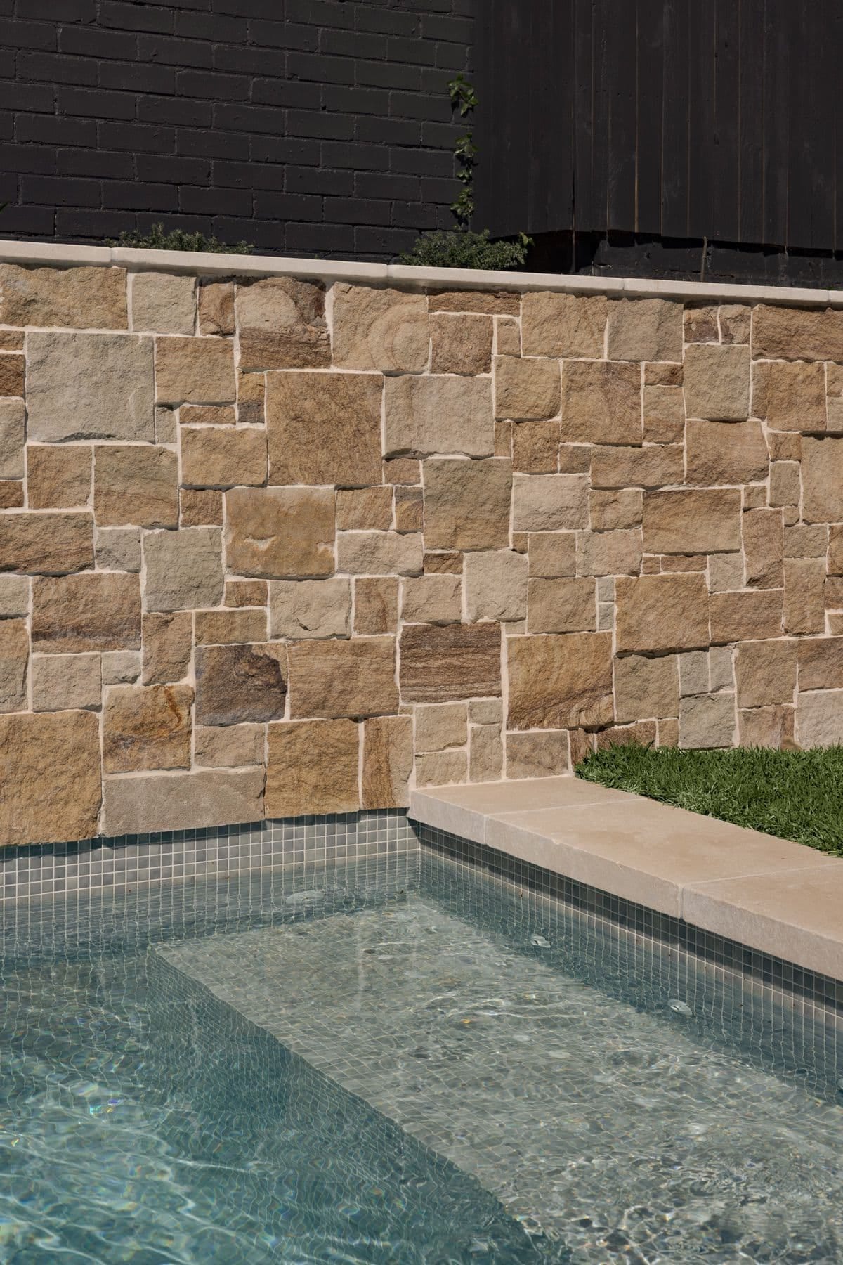 Sandstone block wall meeting the mosaic-tiled waterline, with sandstone coping and lawn at the pool edge.