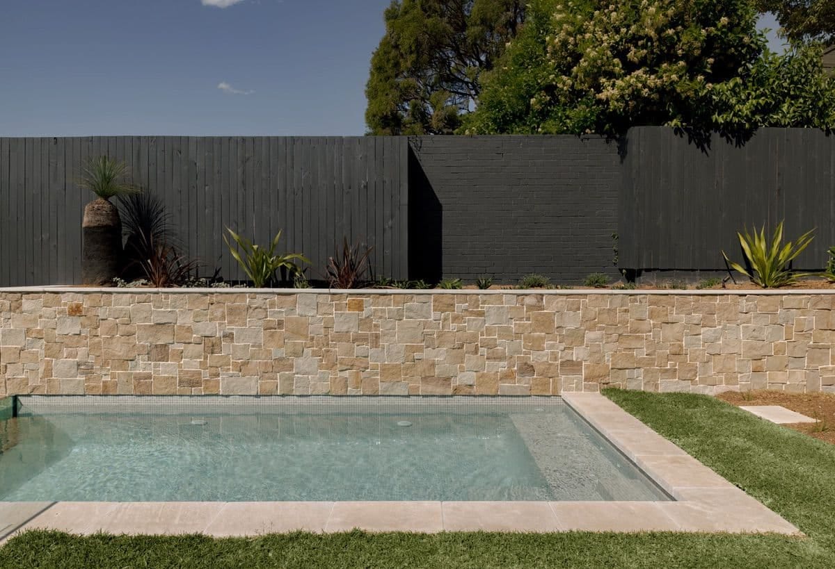 The pool from the lawn, a low sandstone retaining wall behind it and grass trees planted along a dark timber fence.