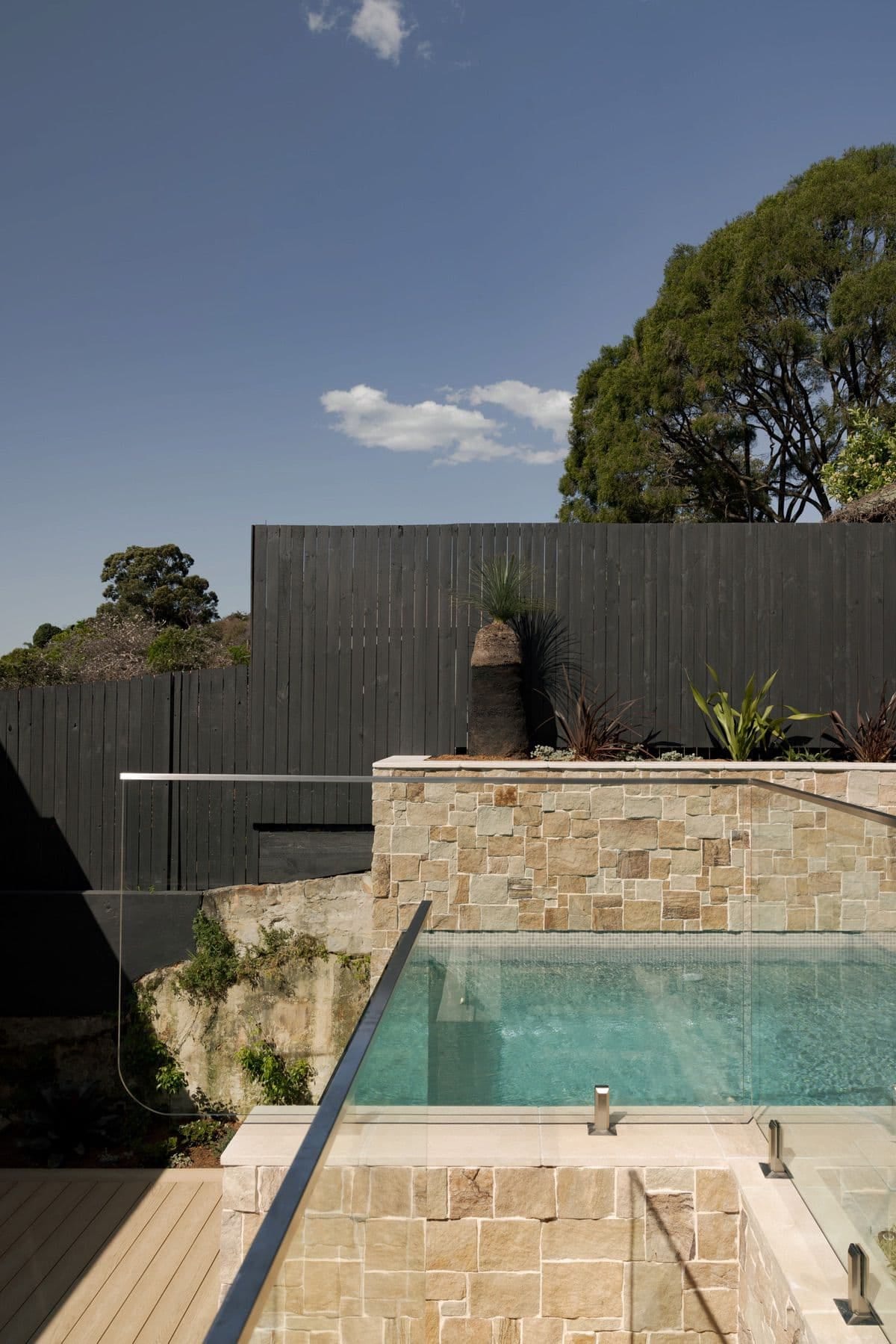 Frameless glass pool fencing above a sandstone wall, with the original rock outcrop exposed below and a grass tree beyond.