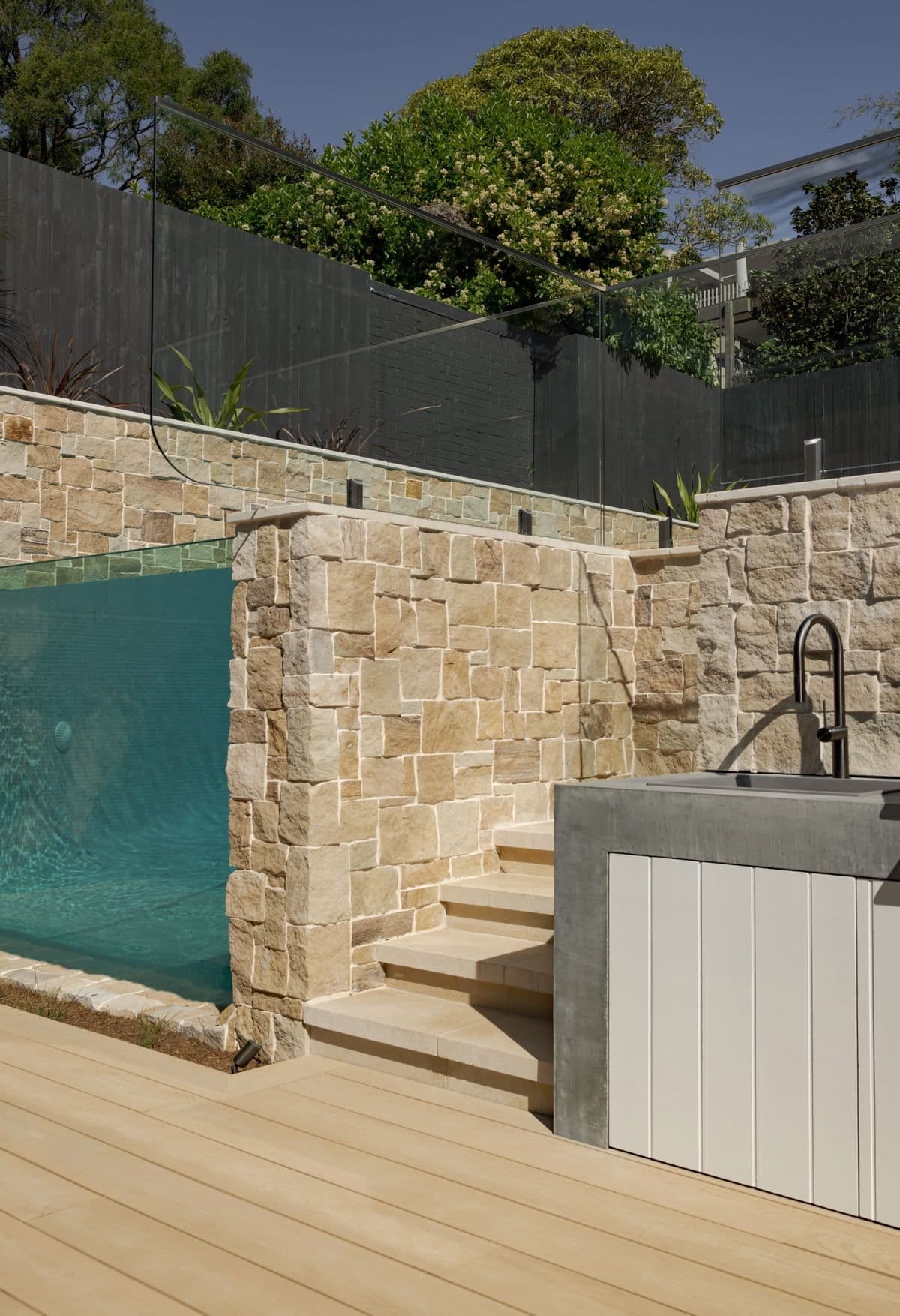 The edge of the glass viewing wall beside sandstone steps rising to the outdoor kitchen and its black tapware.