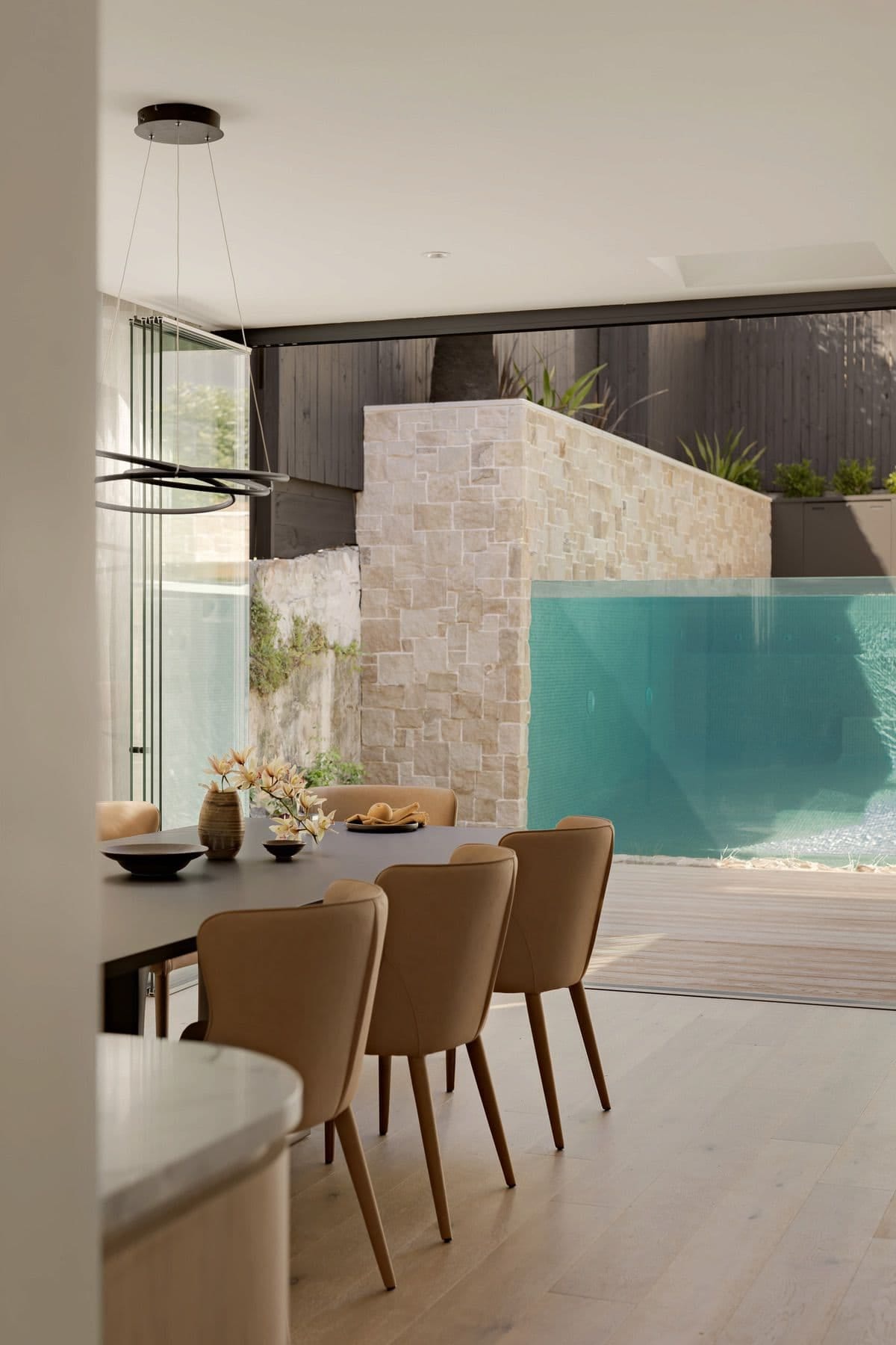 The dining table and tan leather chairs, with the glass wall of the pool filling the opening beyond.