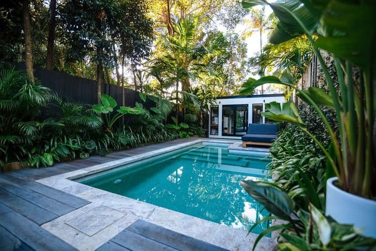 The pool from the deck, palms and tropical planting crowding the black fence, a daybed outside the studio.