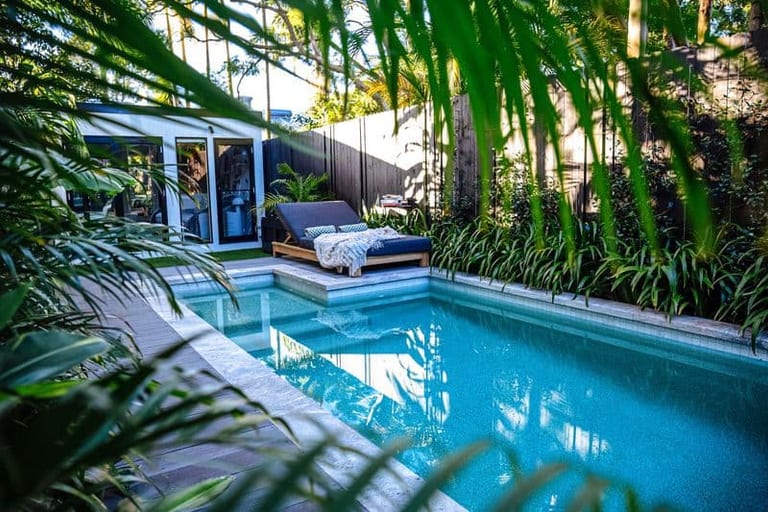 The pool seen through overhanging palm fronds, the daybed and lit studio at the far end.