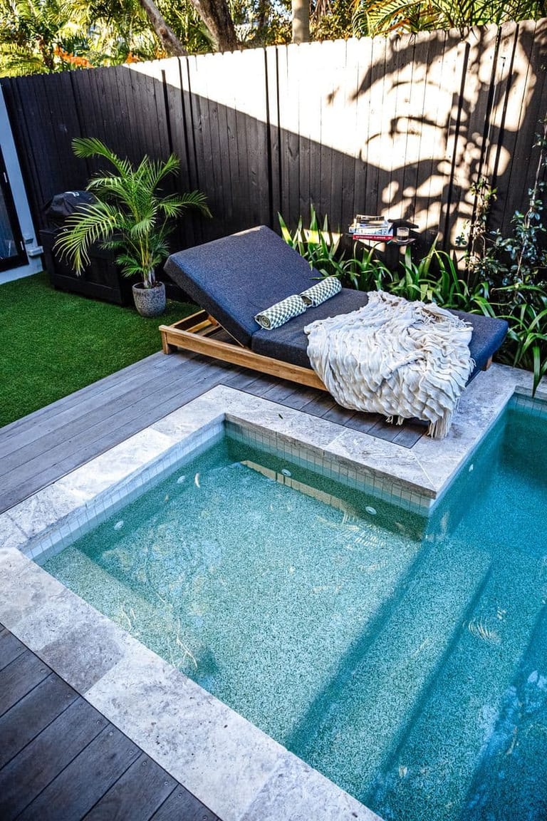 A timber daybed with cushions and a throw on the deck beside the pool's tiled edge.