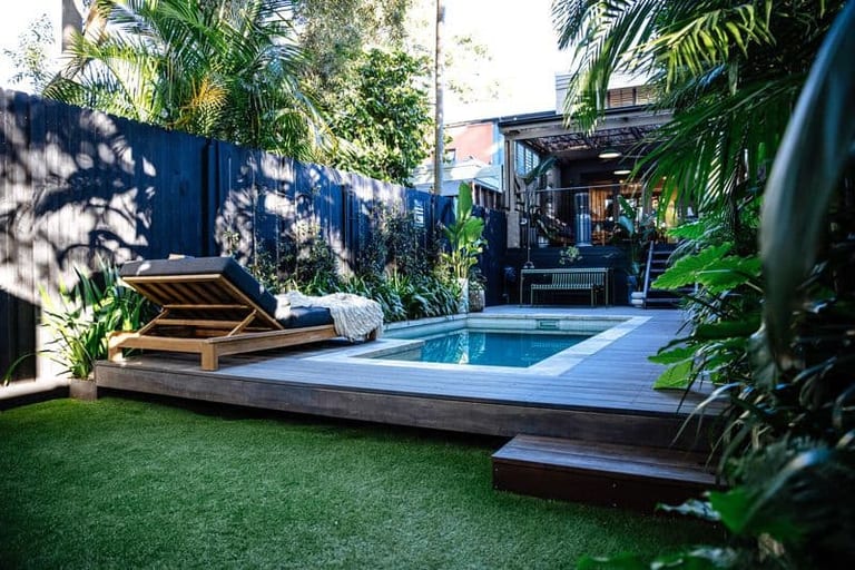 A timber lounger on the deck beside the pool, with lawn in the foreground and the house beyond.