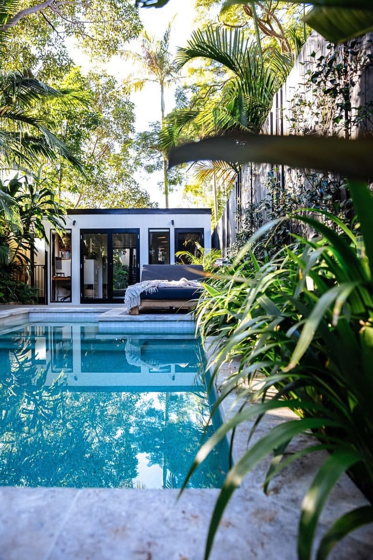 The pool surface reflecting palms, with the studio and daybed seen past foreground foliage.