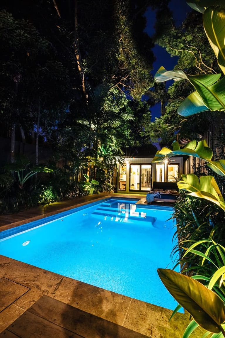 The lit pool at night framed by tropical foliage, the studio's doors open and warm inside.