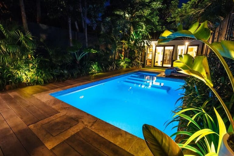 The pool lit at night from the deck, planting uplit along the fence and the studio glowing.