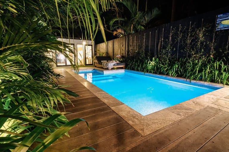 The pool and travertine deck at night, with uplighting through the palms and the studio behind.