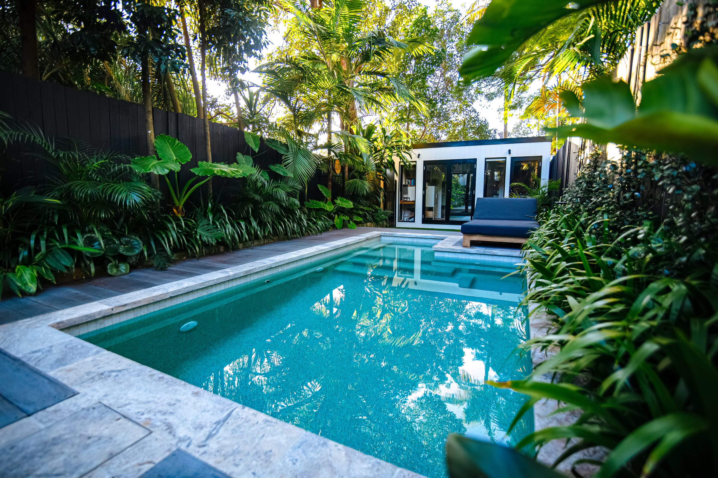 A small tiled pool in a dense tropical courtyard, with travertine coping, a timber deck and a white studio with black-framed doors at the far end.