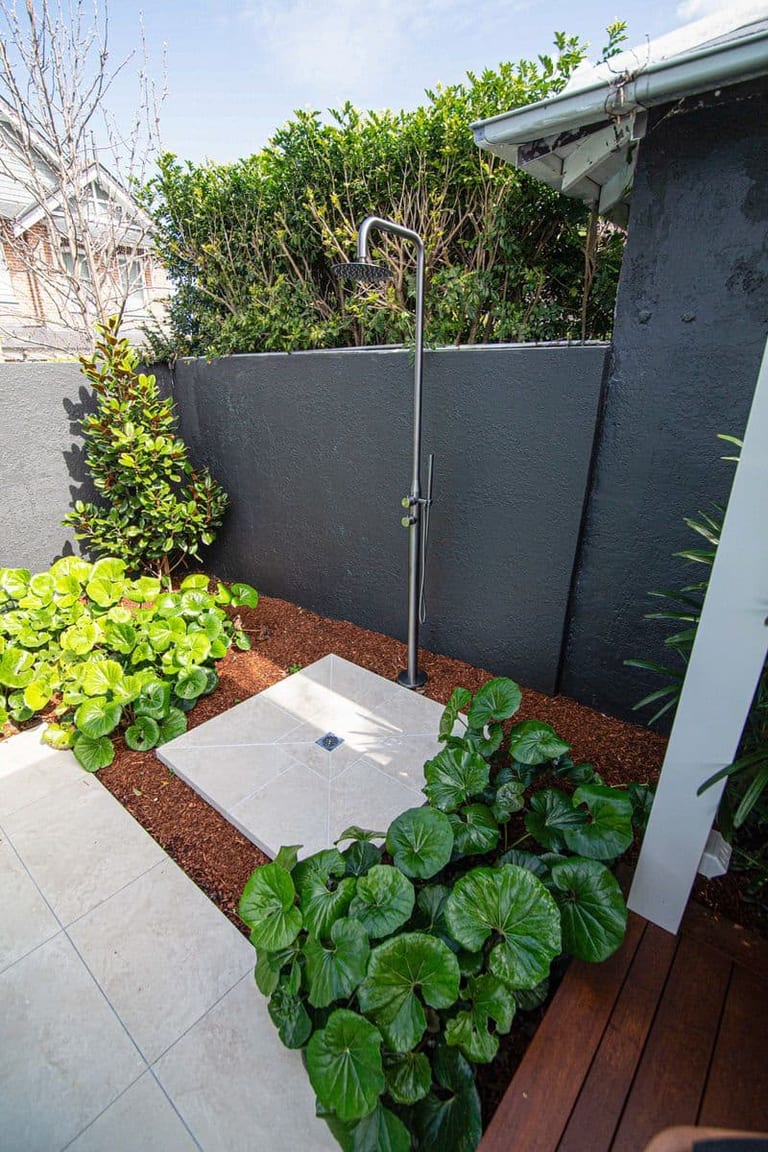 An outdoor shower on a stone pad, set against a dark rendered wall with lush planting.