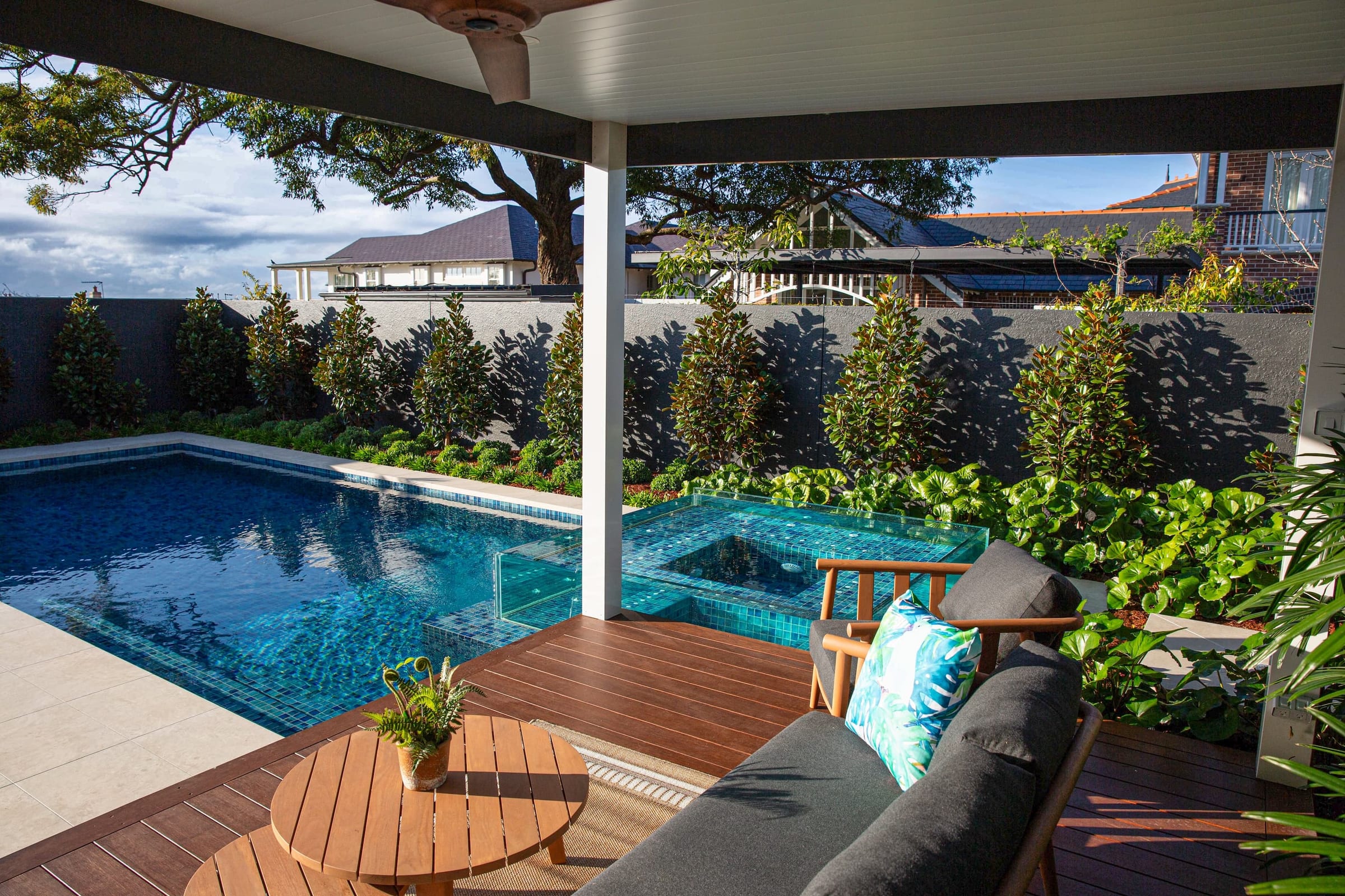 A pool with a raised glass-walled spa, seen from a covered terrace with outdoor lounge seating and a magnolia hedge beyond.