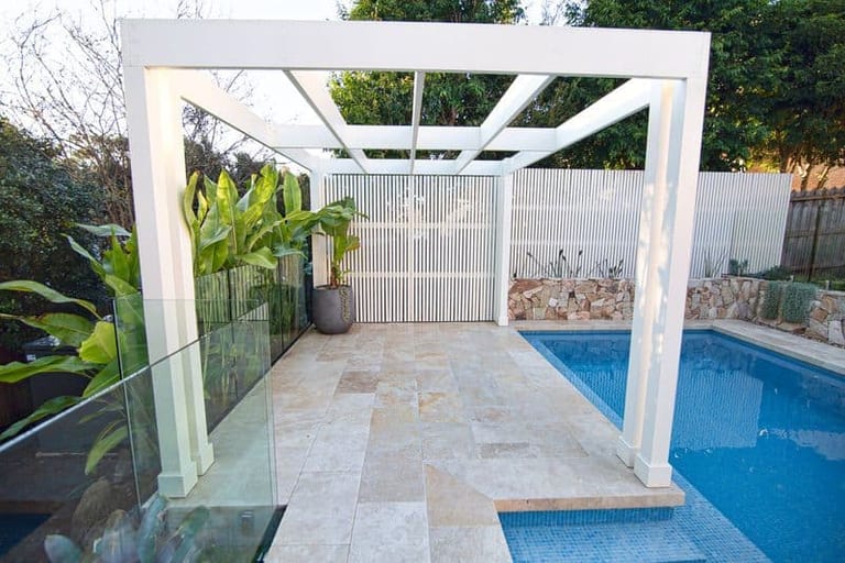 The white pergola over the travertine terrace, its slatted screen and a potted strelitzia beside the pool.