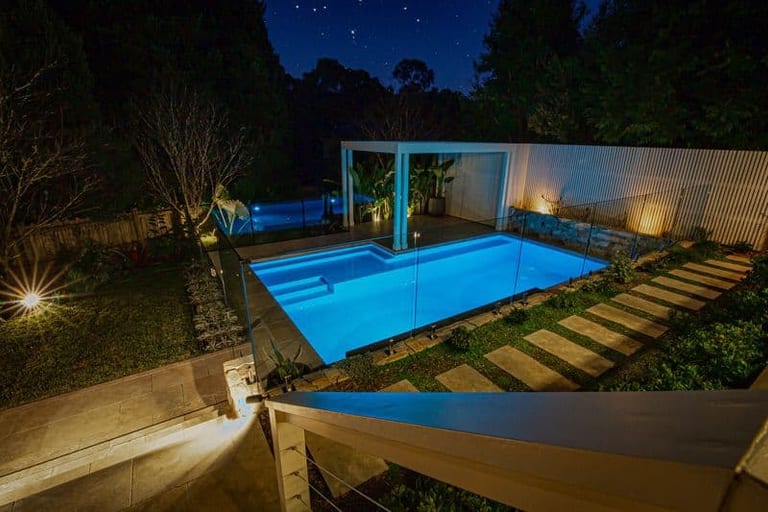 The pool lit blue at night under a star-filled sky, with uplit planting and stepping stones.