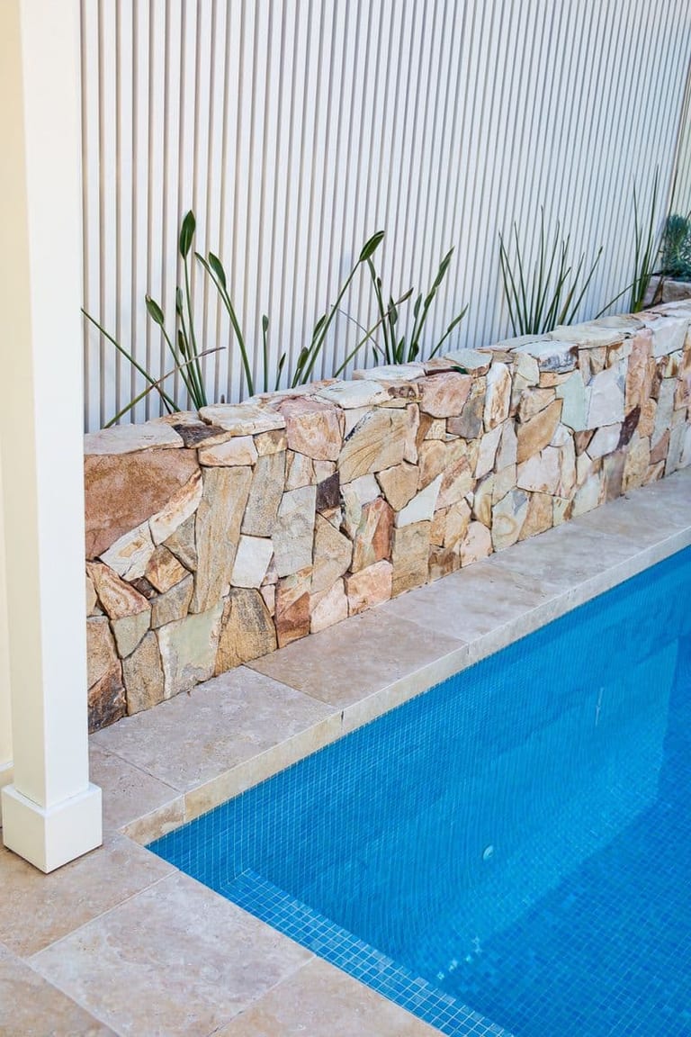 Sandstone retaining wall above the pool's blue tiling, with reeds planted against the white slatted screen.