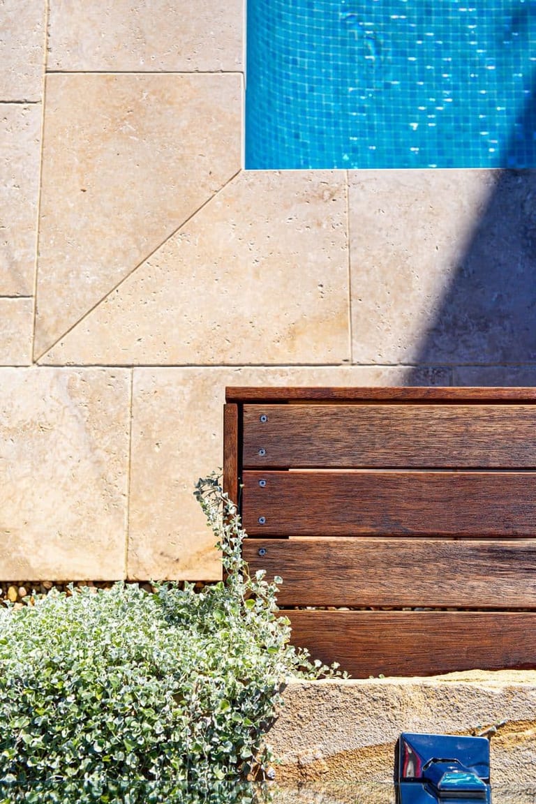 Close detail of travertine paving meeting the pool's blue tiling, with the timber bench and silver groundcover.