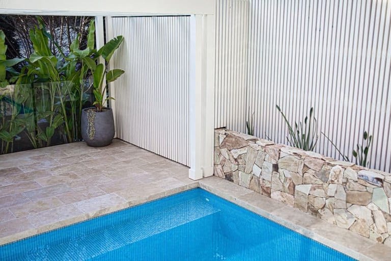 The travertine terrace and slatted screen meeting the sandstone wall at the pool's edge.