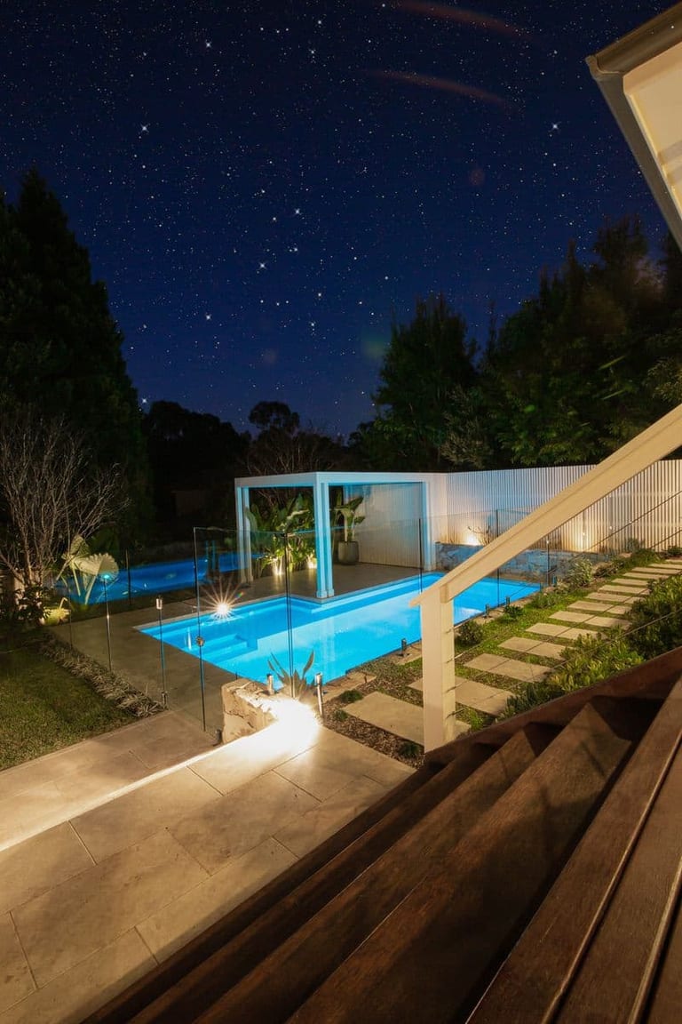 The lit pool at night seen from the house steps, with a star-filled sky above the trees.
