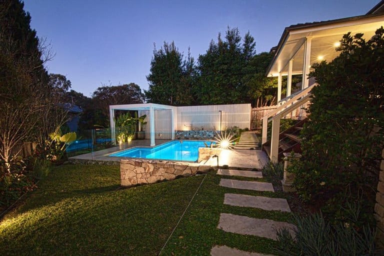 The garden at dusk, stepping stones leading across the lawn to the lit pool and pergola.
