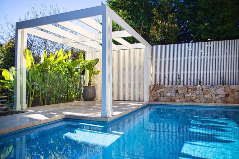 The pergola seen across the pool, its posts and beams framing strelitzia and the slatted screen.