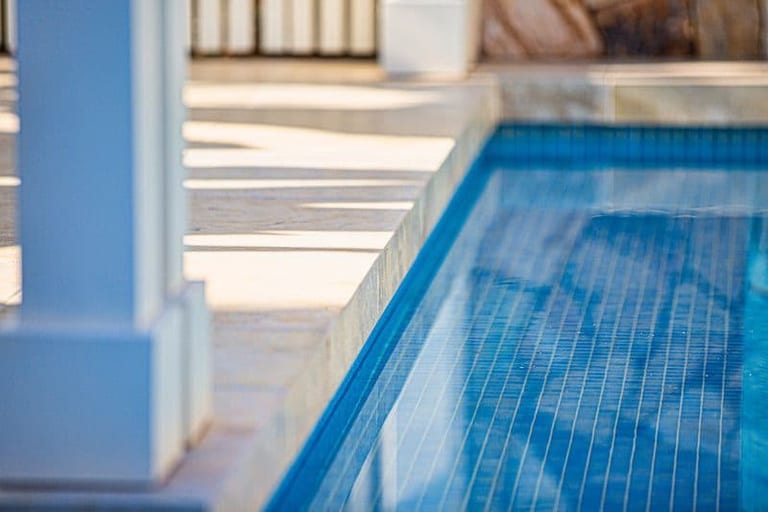 The pool's tiled edge and travertine coping, with a pergola post in soft focus.