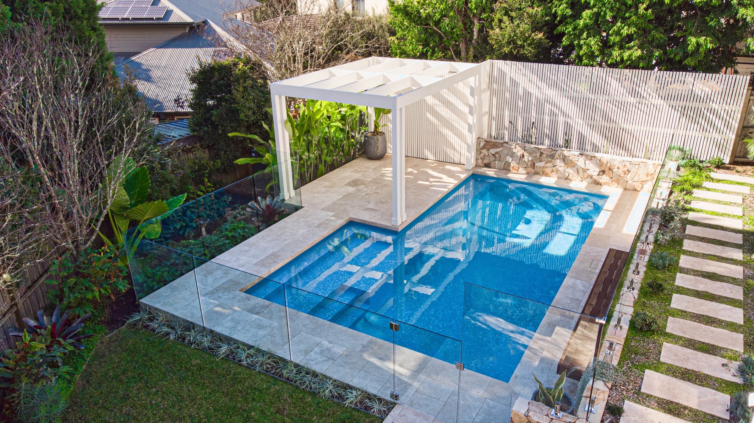 Elevated view of a blue-tiled pool on a terraced block, with a white pergola, slatted screen and sandstone retaining wall behind it.