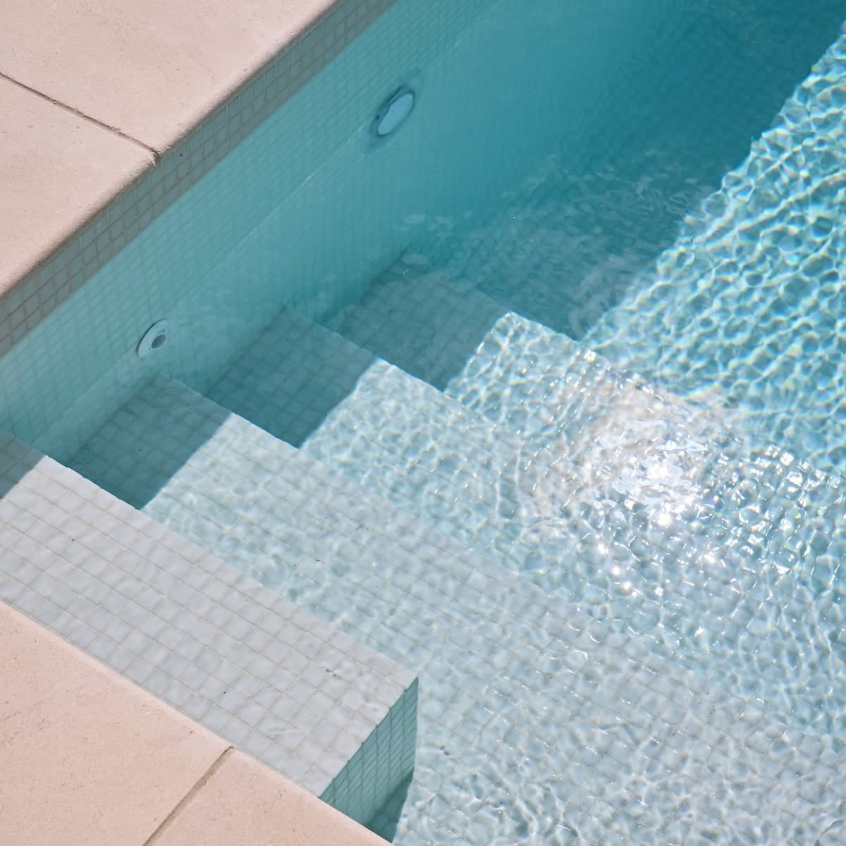 Pale tiled steps leading down into clear water, with a pool light set into the wall and stone coping above.