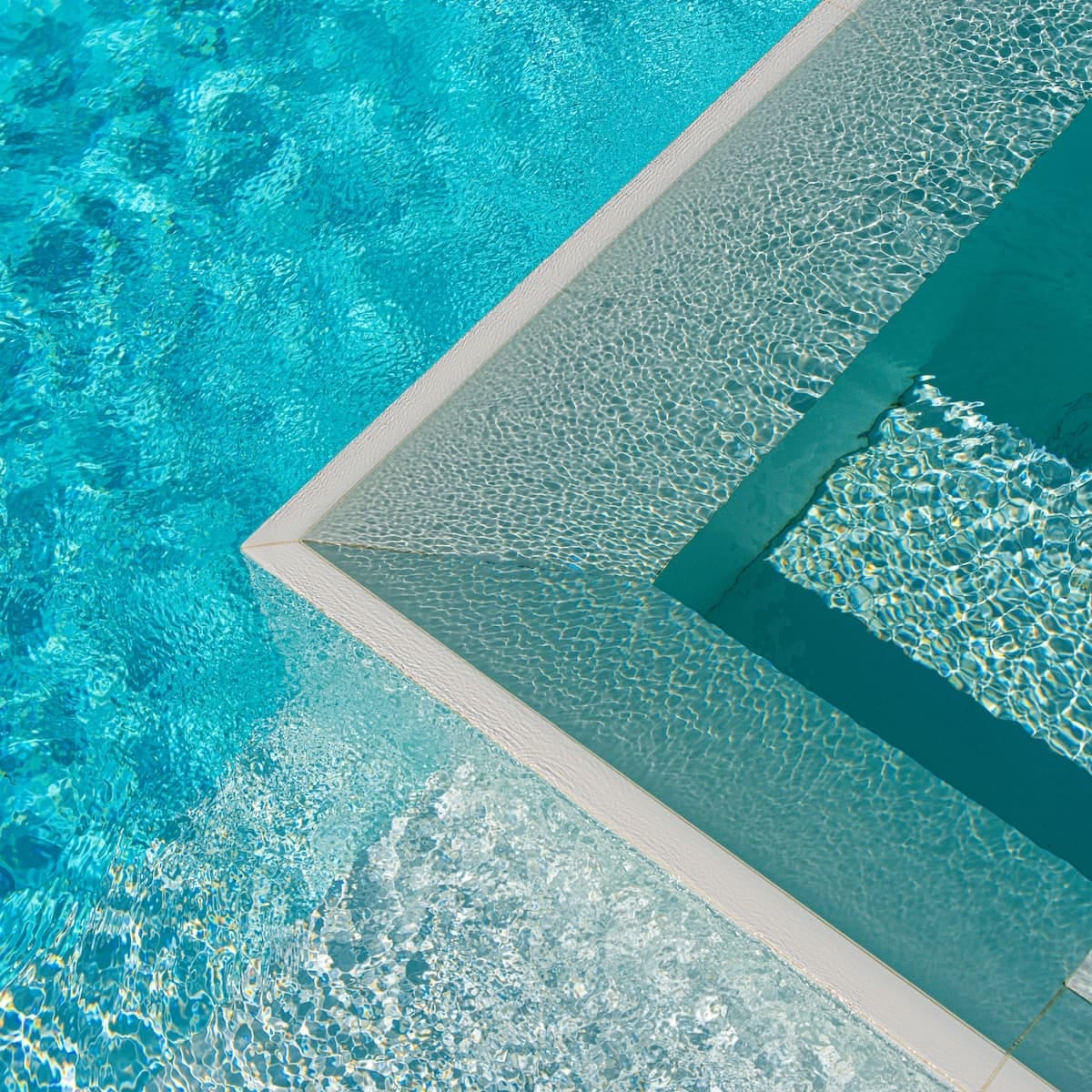 Looking down on the tiled corner of a spa set within a pool, sunlight rippling across the turquoise water.