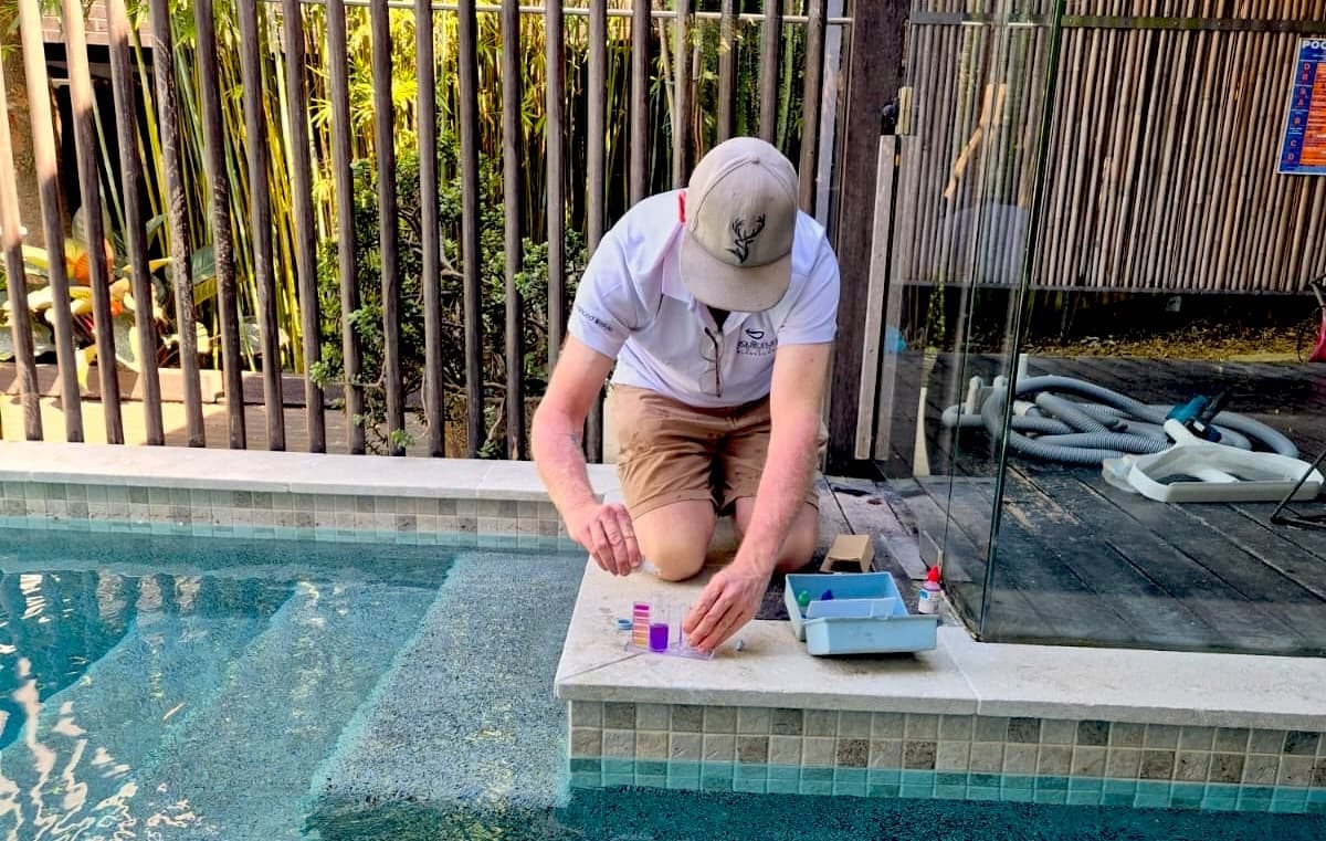 An Equilibrium technician kneeling at the edge of a tiled pool, testing a water sample with a test kit.